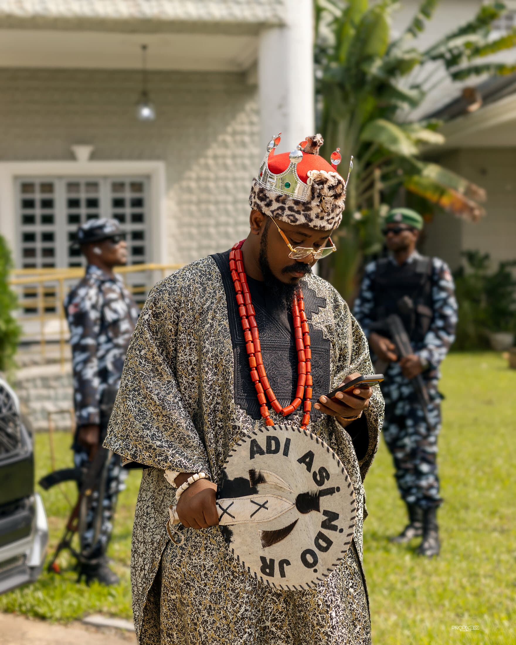 ABJKOREA (Victor) in royal chieftaincy regalia — beaded crown, coral and hand-tooled staff — standing before his estate.